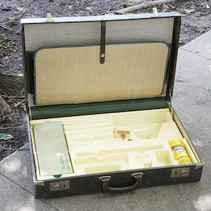 May include: A dark brown rectangular art supply case with a hinged lid and metal latches. The interior features a cream-colored molded plastic tray with designated compartments, containing a green box labeled OIL COLORS and a yellow bottle of thinner.
