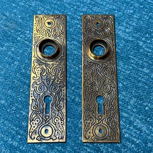 May include: Two antique brass door plates with ornate, symmetrical scrollwork and keyhole cutouts. Each plate features a circular opening for a doorknob and a decorative border. The plates are rectangular and show signs of age.