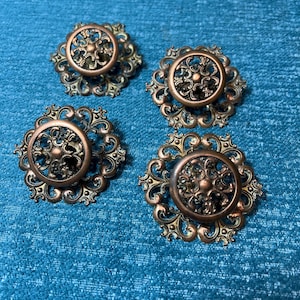 May include: Four antique bronze-colored cabinet knobs with ornate, floral-inspired designs. Each knob features a central circular element surrounded by intricate, swirling patterns. The knobs are set against a textured, teal-colored background.
