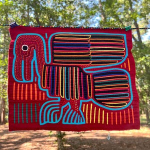 May include: A red textile with a bird design. The bird is outlined in turquoise, with black, yellow, orange, and blue stripes. The bird's eye is a white circle. The textile is hanging on a clothesline with clothespins.