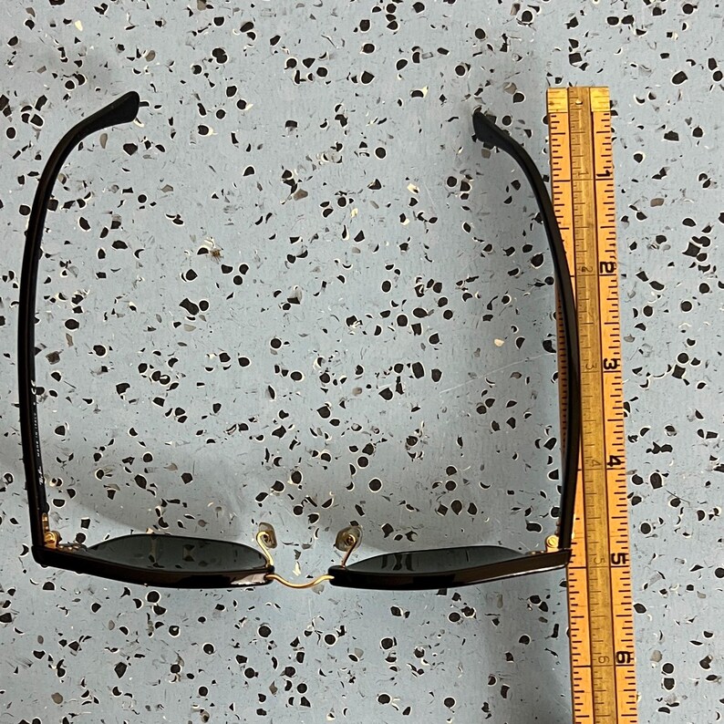 Op de afbeelding: Zwarte zonnebril met een gouden brug en zwarte monturen. De zonnebril is ongeveer 14 cm lang, gemeten met een houten liniaal. De glazen zijn donker en de pootjes zijn licht gebogen.