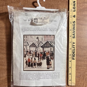May include: A close-up of a needlepoint embroidery kit featuring a black and white illustration of a bustling market scene. The kit includes Appleton crewel wool, a needle, and instructions. The finished piece will be approximately 12 inches by 13 inches.