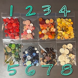 May include: Eight bags of assorted buttons, numbered 1 through 8. Each bag contains a variety of colors, shapes, and sizes of buttons. The buttons are arranged in a grid pattern on a gray surface.