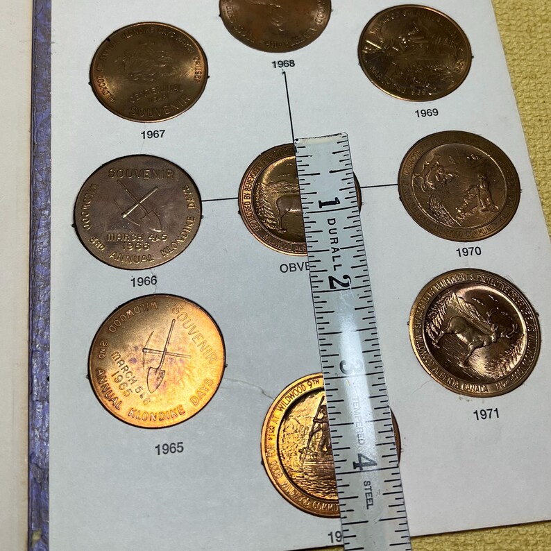 May include: A collection of vintage copper coins, each approximately 1 inch in diameter, displayed in a coin album. The coins are dated from 1965 to 1971, with various designs and text inscriptions. A ruler is placed over the coins.