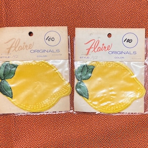 May include: Two yellow lemon-shaped iron-on patches with green leaves. The patches are packaged in clear plastic bags with white cardstock backing. The cardstock has the text "Flaire Originals" and "1/00" printed on it.