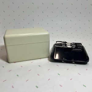 May include: An off-white, rectangular box with a hinged lid sits next to a black address book. The address book has alphabetical tabs and a curved design. The background is a light pink and white pattern.
