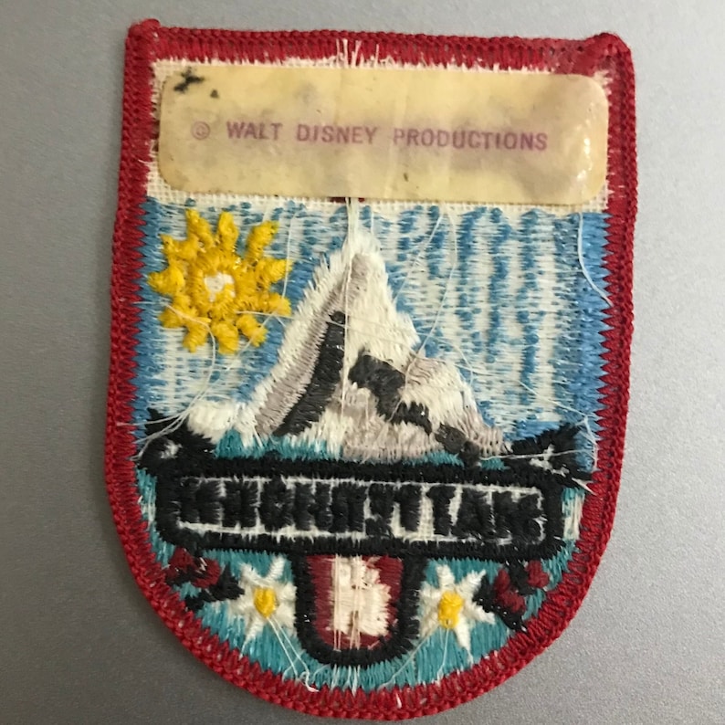 Puede incluir: Un parche bordado vintage que presenta una escena de monta&ntilde;a con un sol, un pico de monta&ntilde;a blanco y el texto "Neuschwanstein" en negro. El parche tiene un borde rojo y un fondo azul con costuras blancas. La parte inferior del parche presenta una bandera suiza roja y blanca con flores blancas.