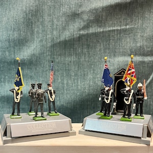 May include: A set of two toy soldier displays, each featuring a group of five toy soldiers in uniform holding flags. The flags are American, British, and a yellow and blue flag. The displays are on a black base with the words "COLLECTION" and "Manufactured in London England".
