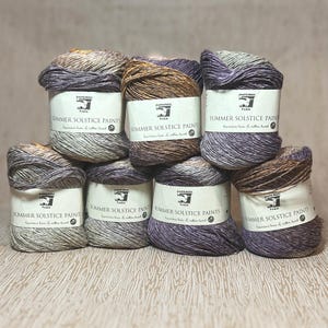 May include: A collection of yarn skeins in various shades of purple, brown, and gray. Each skein has a white label with the text "SUMMER SOLSTICE PAINTS". The yarn is arranged in a stacked display, showcasing the colors and textures.