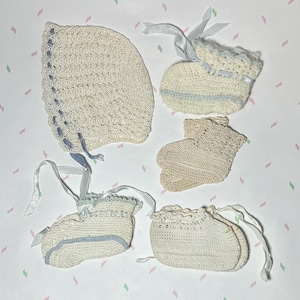 May include: A collection of handmade baby items, including a bonnet and three pairs of booties. The items are crocheted in off-white yarn with light blue accents and ribbons. The booties and bonnet are arranged on a patterned surface.