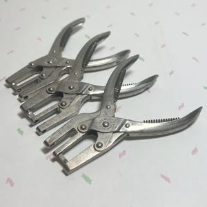 May include: Four vintage silver-toned metal hole punch pliers. The pliers have a curved handle with a textured grip and a rectangular punch head. The tools are arranged diagonally, with the handles open.