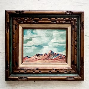 May include: A framed painting of a desert landscape with a mountain range in the background and a blue sky with white clouds. The frame is dark brown with a carved design.