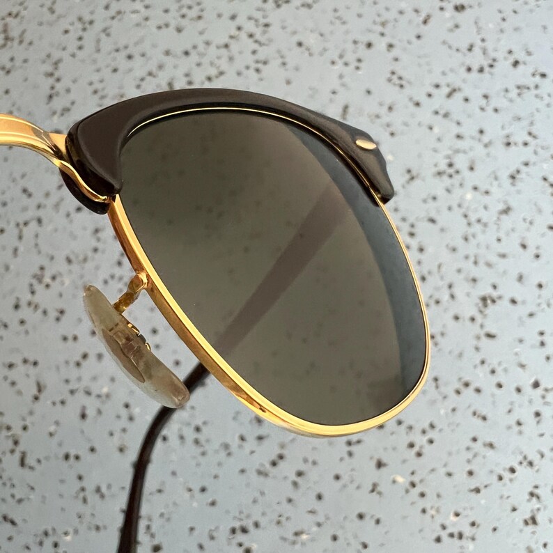 Op de afbeelding: Close-up van een zonnebril met een goudkleurig montuur en donkere glazen. De bovenkant van het montuur is zwart en de neuspads zijn transparant. De zonnebril heeft een klassiek, tijdloos ontwerp.