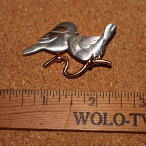 May include: A silver and gold tone brooch featuring two birds perched on a branch. The birds are facing each other and appear to be kissing.