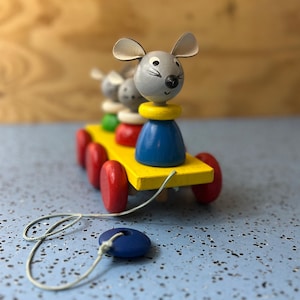 May include: A wooden pull toy featuring a family of mice riding on a yellow cart with red wheels. The mice are gray with white ears and black eyes. The cart has a blue pull-along disc attached.