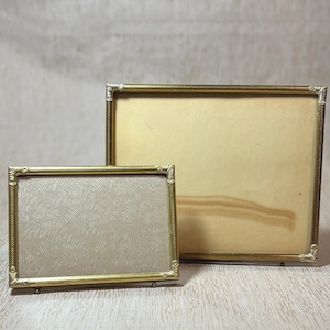May include: Two vintage gold-toned picture frames. The smaller frame has a light-colored patterned backing, while the larger frame has a plain, off-white backing. Both frames have ornate corner detailing and a rectangular shape.
