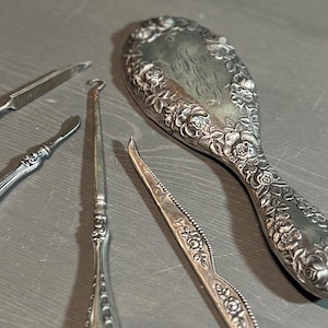 May include: A set of antique silver-plated grooming tools, including a hand mirror, a nail file, a cuticle pusher, and a shoe horn. The mirror has a floral design and is engraved with the initials "M.E."