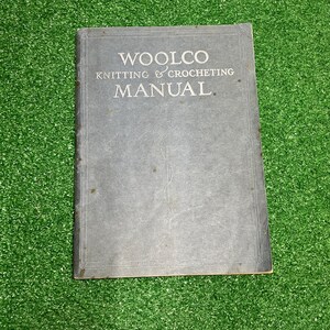 May include: A rectangular gray manual titled WOOLCO KNITTING & CROCHETING MANUAL in white text. The cover features a central line-art illustration of a ball of yarn with a single strand extending downward and a decorative border frame around the edges.