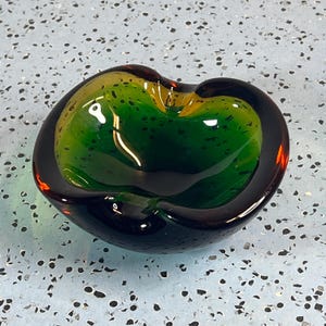 May include: A thick-walled, rounded glass bowl featuring a deep emerald green interior that transitions into a dark, near-black outer rim. The glass has a smooth, polished surface with a heavy, organic shape and a wide, shallow basin design.