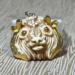 May include: A detailed pendant featuring a stylized yak head. The pendant has a cream-colored face with gold-colored accents on the horns and edges. The yak's face has detailed features, including eyes, a nose, and a mouth. The pendant is designed to be worn as jewelry.