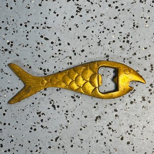 May include: A gold-toned metal bottle opener shaped like a fish. The object features a textured scale pattern along its body, a pointed tail, and a circular eye near the head. The bottle opening mechanism is integrated into the midsection of the fish.