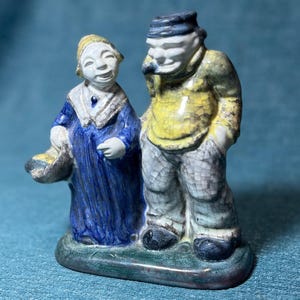 May include: A ceramic figurine depicting a man and woman in traditional attire. The woman wears a blue dress and holds a basket. The man is in a yellow jacket and gray pants. The base is green and the figurine has a crackle glaze.