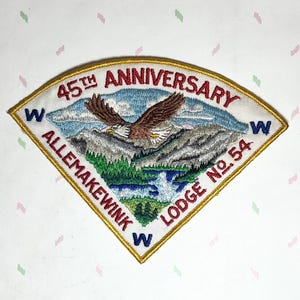 May include: Embroidered patch with a gold border, featuring an eagle in flight over a mountain landscape. Text includes "45th Anniversary," "Allemakeewink Lodge No. 54," and "W" on a white background.