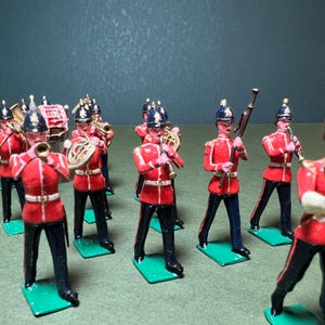 May include: A collection of toy soldiers in a marching band formation. The figures are dressed in red jackets, black pants, and blue helmets. Some soldiers hold musical instruments, while others carry rifles. The figures stand on green bases.
