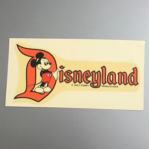 Puede incluir: Pegatina vintage de Disneyland con Mickey Mouse con un atuendo rojo y negro, de pie frente a una "D" naranja estilizada con la palabra "Disneyland" en letras naranjas. La pegatina incluye la informaci&oacute;n de copyright "&copy; WALT DISNEY PRODUCTIONS".