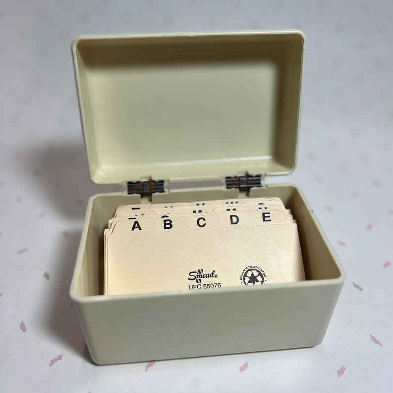 May include: An open, off-white address card box with a hinged lid. The box contains a stack of cream-colored index cards labeled A, B, C, D, and E. The brand name "Smead" is visible on the cards.