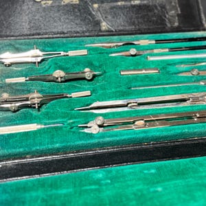 May include: A collection of vintage drafting tools, including compasses and dividers, neatly arranged in a black case with a green velvet lining. The tools are made of silver-colored metal and are designed for precision drawing and measurement.