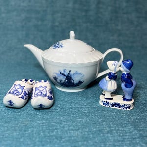 May include: A white ceramic teapot with blue floral and windmill designs, a pair of miniature blue and white clogs, and a figurine of two kissing figures. The items are arranged on a blue textured surface.