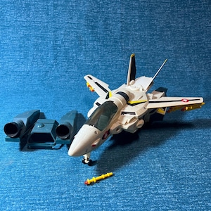 May include: A white and black toy fighter jet with yellow and red accents, and a clear cockpit. The jet has detachable parts and is accompanied by two blue-grey engine components and a yellow missile.