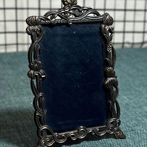 May include: Ornate, antique-style picture frame with a dark blue velvet backing. The frame is made of a dark metal with intricate floral and scrollwork detailing. It has a small easel back for tabletop display.