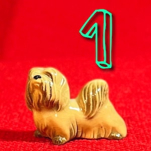 May include: A small, beige ceramic figurine of a dog with its head tilted up. The dog has a short, fluffy coat and a long tail. The figurine is sitting on a red surface.