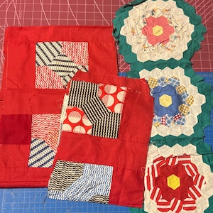 May include: A collection of handmade quilt blocks in various stages of completion. The blocks feature red fabric with geometric patterns in black, white, and blue. Several finished floral-shaped quilt pieces are also present, showcasing a mix of colors.