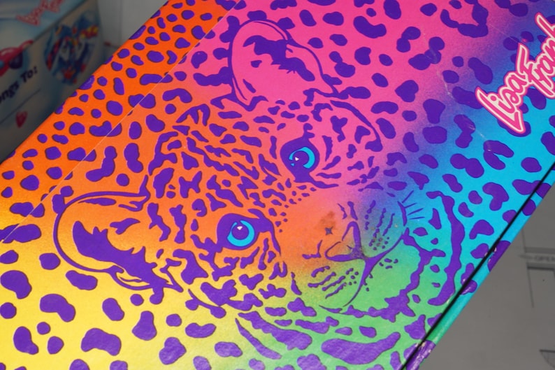 May include: A rectangular box featuring a vibrant, multi-colored leopard print pattern in shades of orange, pink, purple, and blue. The face of a leopard is centered on the surface, and the text Lisa Frank is printed in a white script font.