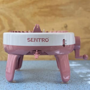 May include: A pink and white Sentro knitting machine with a hand crank and four legs. The machine has the word "SENTRO" printed on the side. The machine is designed for circular knitting projects.