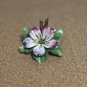 May include: A small, hand-painted porcelain flower with purple, white, and green accents. The flower is on a brown background.