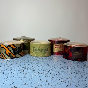 May include: Five vintage Faberge round containers with clear lids. The containers have various patterns and colors, including orange and black, green, red, and gold. Each container has the Faberge logo.