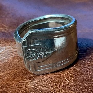 May include: A silver spoon ring with a floral design. The ring is made from a bent spoon and has a wide band.