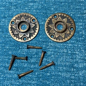 May include: Two antique brass door rosettes with a floral design, along with several matching screws. The rosettes have a circular shape with a central hole and three smaller holes around the perimeter. The screws are a dark bronze color.