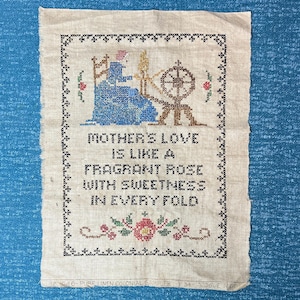 May include: A vintage cross-stitch sampler on linen. The design features a woman at a spinning wheel, with the text "MOTHER'S LOVE IS LIKE A FRAGRANT ROSE WITH SWEETNESS IN EVERY FOLD." The border is black, with small floral accents.