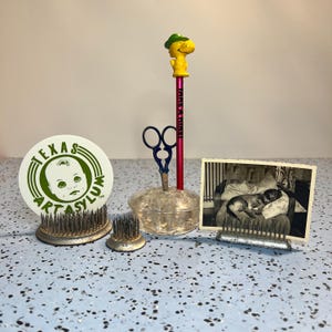 May include: A collection of vintage metal flower frogs, including one holding a round white sign with green text reading TEXAS ART ASYLUM, a clear glass dish with blue scissors and a yellow pencil topper, and a black and white dog photo.