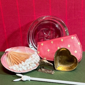 May include: A set of decorative items including a glass ashtray, a pink leather clutch with a gold fleur-de-lis pattern, a small brass heart-shaped dish, a seashell with pearl-like beads and toothpicks, and a small gold-colored bird figurine.