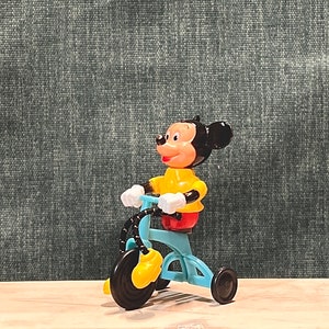 May include: A yellow and black Mickey Mouse figurine riding a blue tricycle with black wheels.