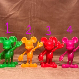 May include: Four plastic Mickey Mouse figurines in different colors: green, orange, red, and pink. Each figurine is standing with one arm raised and the other arm at its side.