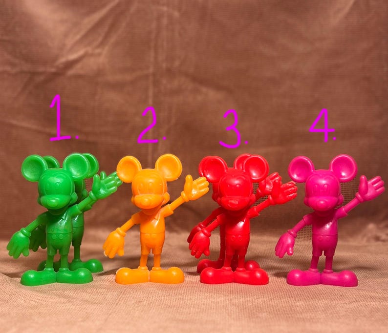 May include: Four plastic Mickey Mouse figurines in different colors: green, orange, red, and pink. Each figurine is standing with one arm raised and the other arm at its side.