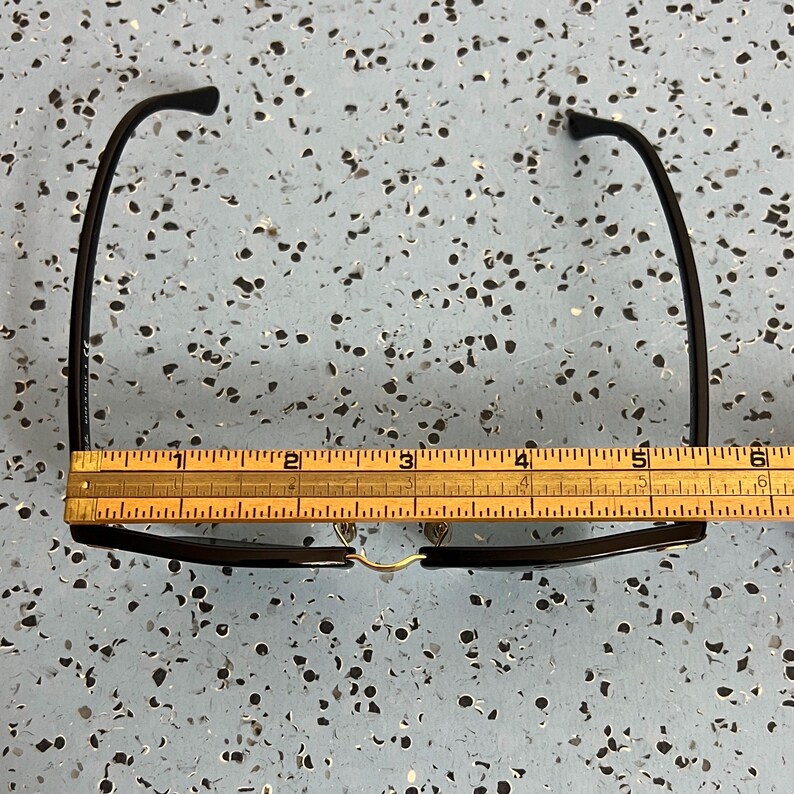 Op de afbeelding: Een zwart montuur bril met een goudkleurige brug wordt gemeten met een houten liniaal. De bril ligt op een gespikkeld blauw oppervlak. De liniaal geeft aan dat de bril ongeveer 15 cm breed is.