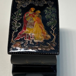 May include: A black wooden box with a hinged lid. The lid is decorated with a painted scene of a couple in a forest setting. The couple is dressed in colorful clothing and the scene is surrounded by a gold border.